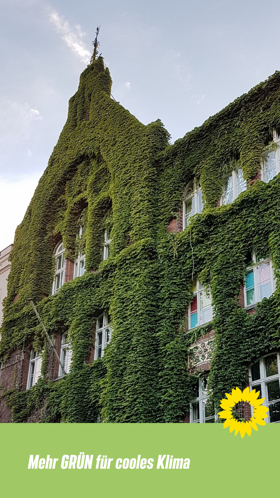 Ein altes Backsteingebäude aus dem 20. Jahrhundert mit großen Fabrikartigen Sprossenfenstern ist überrankt mit Blattgrün. Darüber strahlt hellblaugrauer Himmel.